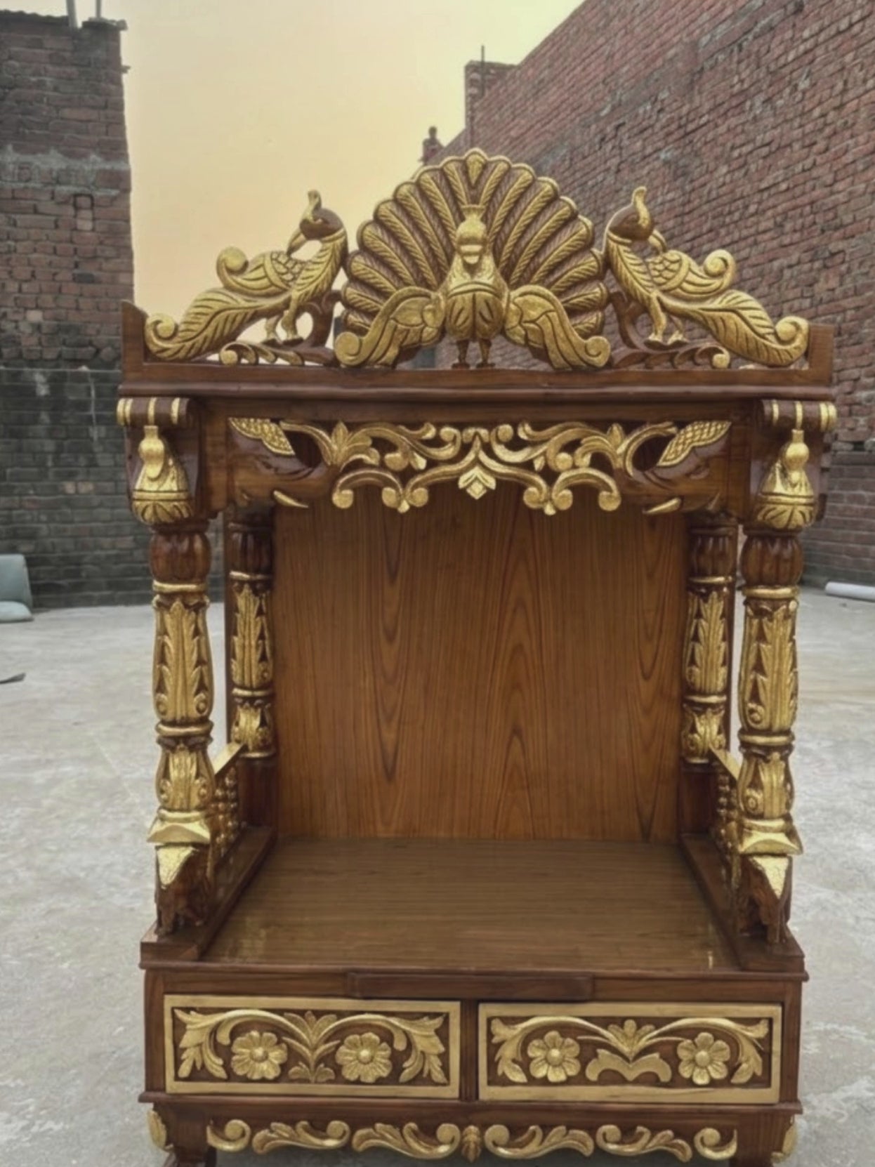 Brown and gold wooden home temple with intricate peacock carving, floral details and carved pillars